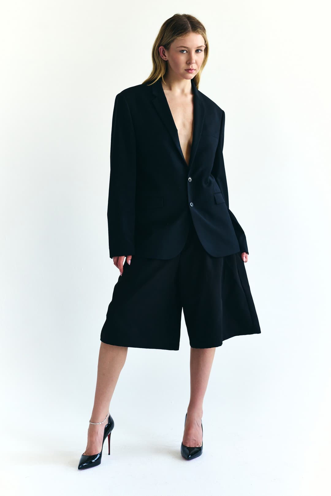 Full-length studio photograph of Sage Burress in a black blazer, tailored shorts, and heels.